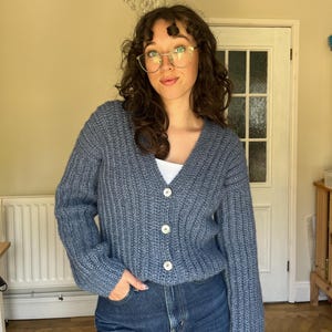 Puede incluir: Un cárdigan azul tejido con cuello en V y tres botones blancos. El cárdigan se combina con unos vaqueros azules. La imagen está tomada en interiores, con una lámpara de araña en el fondo.