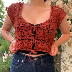 May include: A rust-colored crochet top with a square pattern and a tie at the front. The top is cropped and has short sleeves.
