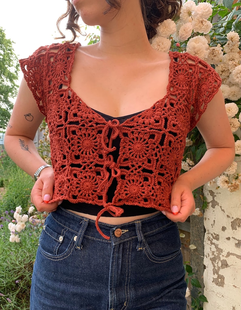 May include: A rust-colored crochet top with a square pattern and a tie at the front. The top is cropped and has short sleeves.