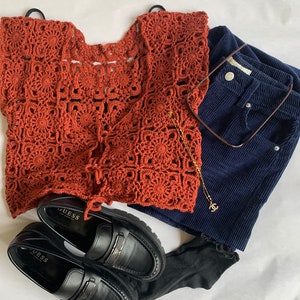 May include: A rust-colored crochet top with a square pattern, black loafers with the word "GUESS" on them, dark blue corduroy shorts, and a gold chain necklace with a small charm.