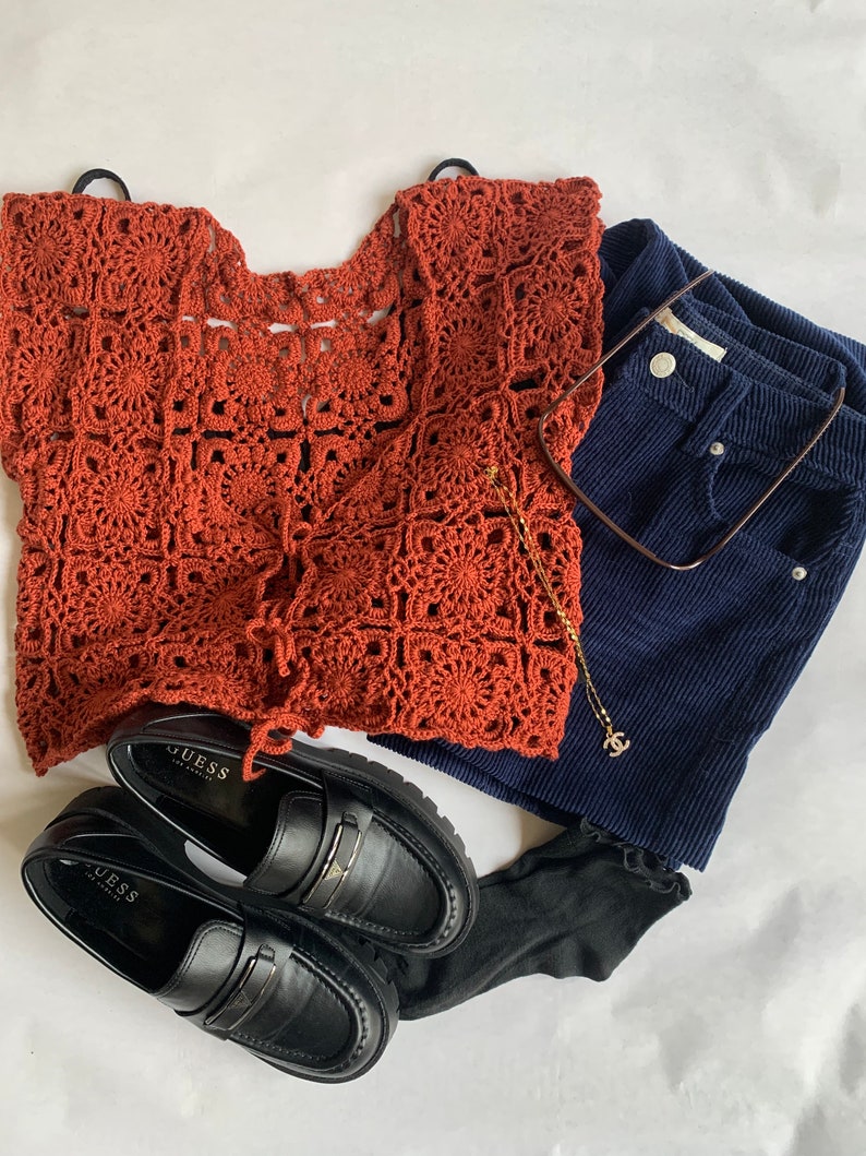 May include: A rust-colored crochet top with a square pattern, black loafers with the word "GUESS" on them, dark blue corduroy shorts, and a gold chain necklace with a small charm.