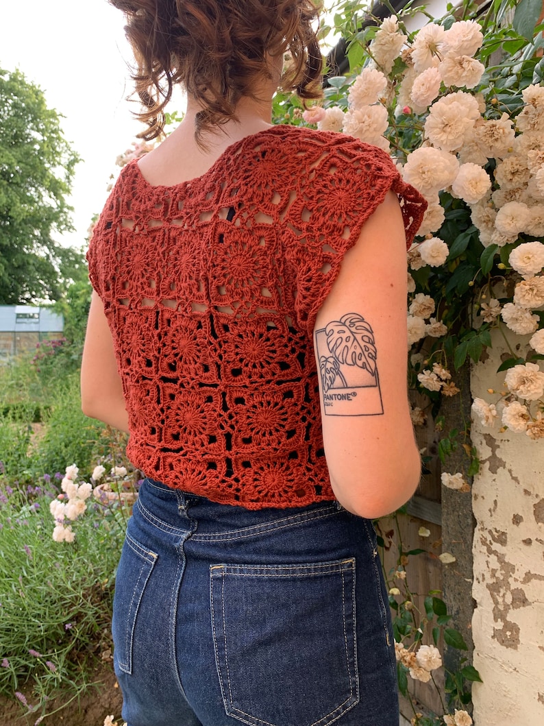 May include: A rust-colored crochet top with a square pattern. The top has short sleeves and a scoop neck. The person wearing the top is standing in front of a bush of white flowers.