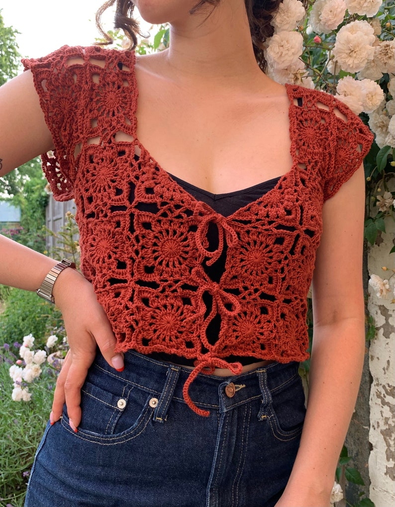 May include: A crocheted top with a tie front and short sleeves. The top is made of a rust-colored yarn and has a square pattern.