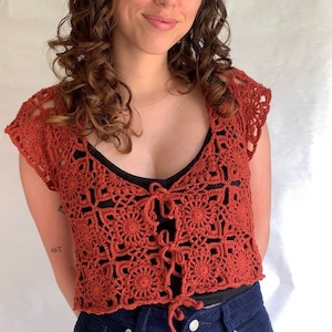 May include: A rust-colored crochet top with a square pattern and a tie at the front. The top is worn over a black tank top and blue jeans.