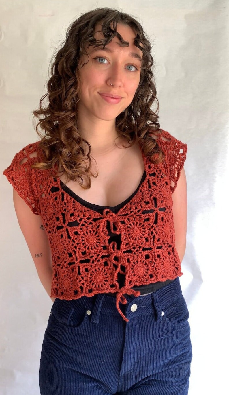 May include: A rust-colored crochet top with a square pattern and a tie at the front. The top is worn over a black tank top and blue jeans.
