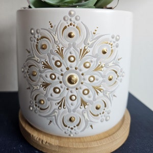 May include: White ceramic plant pot featuring a gold and grey mandala pattern. The pot contains a faux succulent with green and purple leaves. It rests on a round bamboo saucer. A decorative item for home decor.