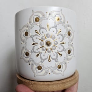 Puede incluir: Maceta de cerámica blanca con un diseño de mandala dorado y gris. La maceta tiene una base de madera.