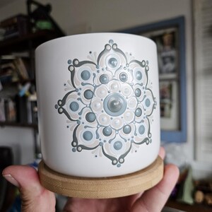Puede incluir: Maceta de cerámica blanca con un diseño de mandala en tonos grises, azules y beige. La maceta descansa sobre un posavasos redondo de bambú. El diseño simétrico es detallado, adecuado para la decoración del hogar. Aproximadamente 10 cm de diámetro.