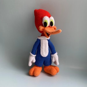 May include: A handmade crochet Woody Woodpecker doll. The cartoon character is depicted with a red head, orange beak and feet, blue body, white gloves and collar. The doll is standing against a light gray background. The text "PDF PATTERN" is on the left side of the image.