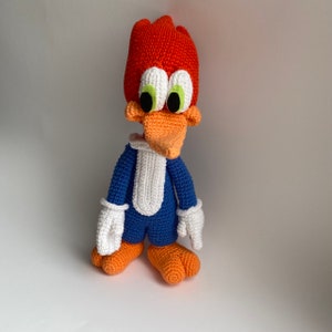May include: A handmade crochet Woody Woodpecker doll. The doll features a red head with a tuft, large white-rimmed eyes with green irises, an orange beak, and blue body with white gloves and a white bib. The feet are orange.