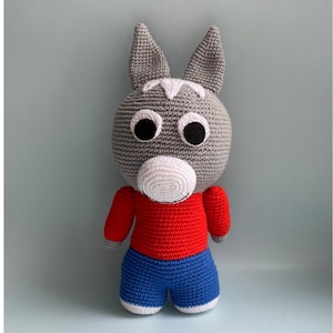 May include: A gray crocheted donkey toy wearing a red shirt and blue pants. The toy has large black eyes and a white nose.