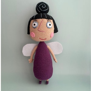 May include: A crocheted fairy doll with purple dress, white wings, and black hair. The doll has a smiling face with blue eyes and pink cheeks.