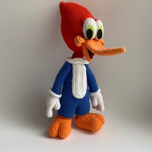May include: A handmade crochet Woody Woodpecker doll. The doll features a red head and beak, blue body, white gloves, and orange feet. The cartoon character is smiling and standing.