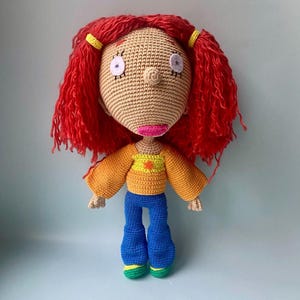 Puede incluir: Muñeca de crochet con pelo rojo, una camisa amarilla y naranja, pantalones vaqueros azules y zapatos verdes.