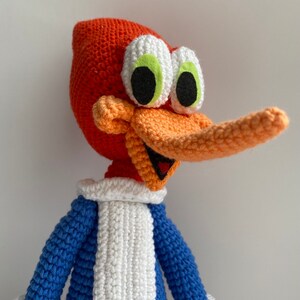 May include: A handmade crochet Woody Woodpecker doll. The doll features a bright orange head and beak, white eyes with black pupils, and a blue and white outfit. The character's signature grin is visible.