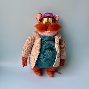 May include: A crocheted plush toy of a cartoon character wearing a pink and purple hat, a peach-coloured jacket, and a teal sweater. The character has orange fur, a red nose, and green eyes.