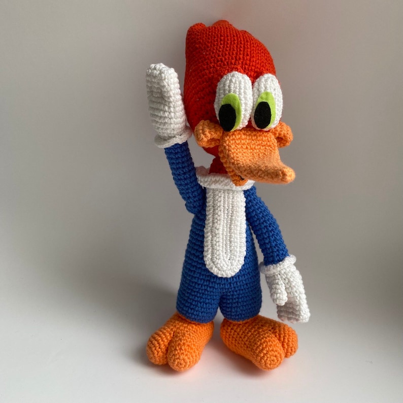 May include: A handmade crochet Woody Woodpecker doll. The cartoon character has a red head, orange beak and feet, blue body, and white gloves. The doll is waving its right hand. The eyes are white with green irises and black pupils.