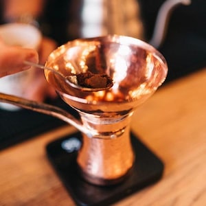 Könnte beinhalten: Eine Kaffeekanne aus Kupfer mit einem trichterförmigen Oberteil, die mit einer Kelle mit Kaffeepulver gefüllt wird. Die Kanne steht auf einer schwarzen Waage.