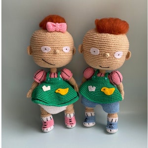 May include: Two crocheted dolls, one with a pink bow and the other with orange hair. They wear green dungarees, blue shorts, and pink and blue shoes. The dolls have white eyes and smiling mouths. The text "PDF PATTERN" is on the left side.