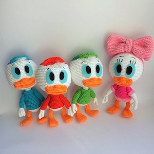 Può includere: Quattro peluche di anatra all'uncinetto in diversi colori. Le anatre sono bianche con becchi arancioni e occhi blu. Indossano magliette di diversi colori: blu, rosso, verde e rosa. L'anatra rosa ha un fiocco rosa sulla testa.
