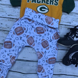 Pode incluir: Leggings brancos para beb&ecirc; com um padr&atilde;o de futebol e X e O. Uma camisa amarela e verde com o texto "GO! PACKERS" e um logotipo "G" verde est&aacute; em cima das leggings.