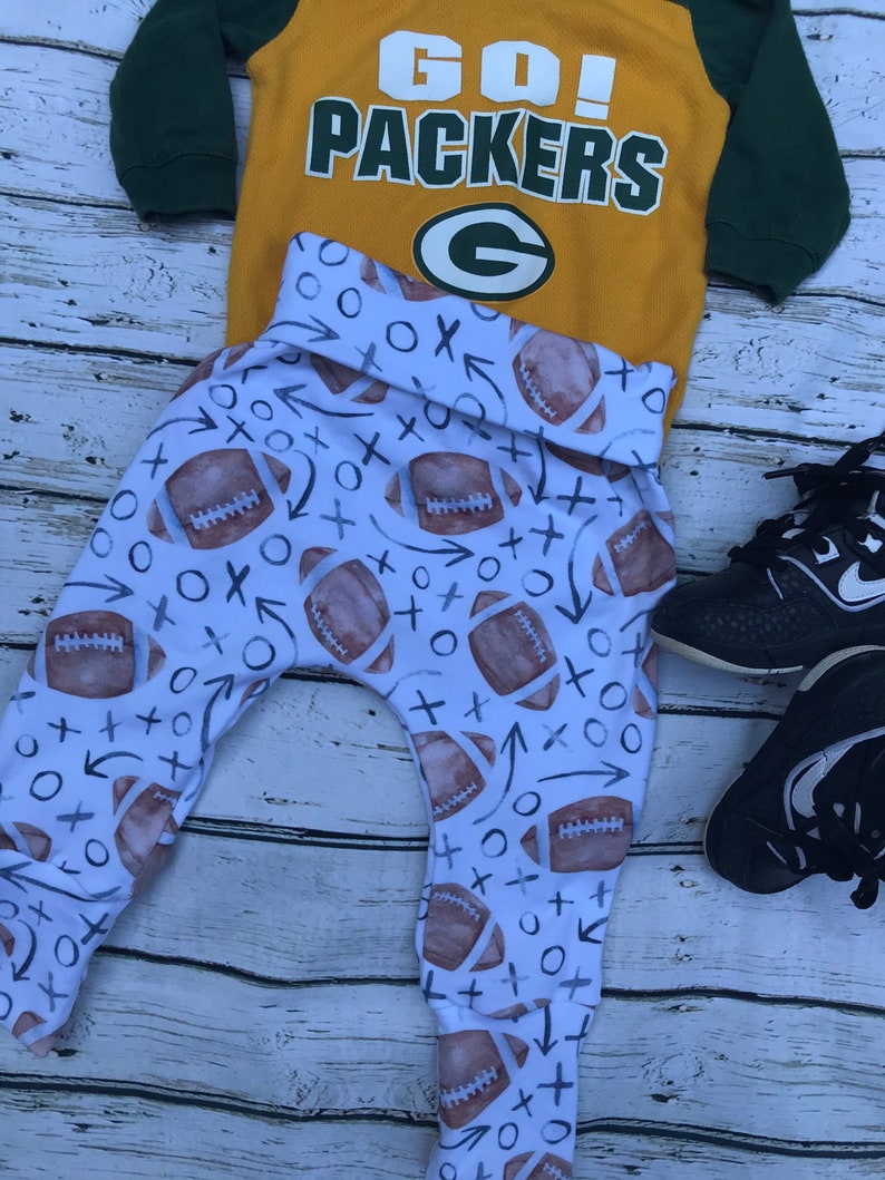 Pode incluir: Leggings brancos para beb&ecirc; com um padr&atilde;o de futebol e X e O. Uma camisa amarela e verde com o texto "GO! PACKERS" e um logotipo "G" verde est&aacute; em cima das leggings.