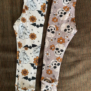 Puede incluir: Dos pares de leggings con temática de Halloween. Un par es blanco con flores naranjas, fantasmas y murciélagos. El otro par es gris con flores naranjas y calaveras.