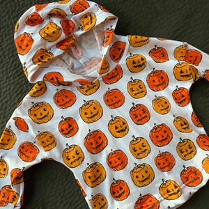 May include: White hooded sweatshirt with a pumpkin pattern. The pumpkins are orange and yellow with black facial features.