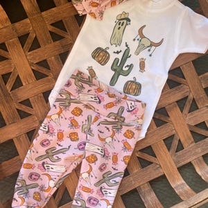 May include: A baby outfit set featuring a white short-sleeved bodysuit with cartoon graphics of a ghost, cactus, and pumpkins. The set includes matching pink leggings and a headband, all with a similar print. The outfit is designed for infants.