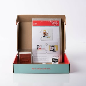 May include: An open cardboard box with a blue and white interior. The box contains a set of scrapbooking supplies, including a paper pad, a stamp, and a stamp pad. The box has the text "Run away with me." printed on the front.