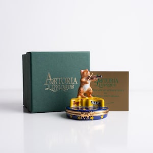 May include: A porcelain figurine of a cat playing a violin, atop a blue and gold box with star and animal designs. The figurine is part of the "Aertoria Limoges" Master Artist Collection, with a green box and certificate.