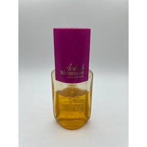Puede incluir: Un frasco de perfume vintage con un cuerpo de vidrio transparente y una tapa fucsia vibrante. El frasco contiene un líquido de color dorado y presenta el texto "Ambush TENDER MUSK SPRAY COLOGNE" en letras doradas.