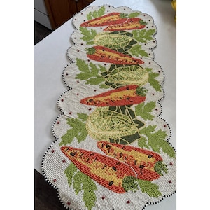 May include: Beaded table runner with a repeating pattern of red and yellow peppers and green gourds with green leaves. The runner has a scalloped edge with black and white beads. The background is a light beige.