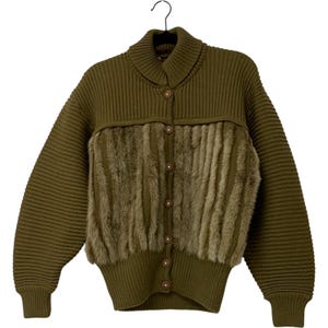 Op de afbeelding: Olijfgroen vest met een geribbelde gebreide design en een imitatiebont paneel aan de voorkant. De trui heeft een knoopsluiting en een kraag. De mouwen zijn lang en de trui hangt aan een zwarte hanger.