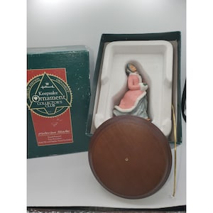 Peut inclure: Une figurine en porcelaine d'une femme vêtue d'une robe rose et d'une fourrure blanche, tenant un bouquet de fleurs. La figurine fait partie d'une édition limitée du Hallmark Keepsake Ornament Collector's Club. La figurine est présentée sur un support en bois.