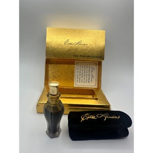 Op de afbeelding: Een Estée Lauder Skin Perfume Flacon set. De gouden doos is open en onthult een kleine parfumfles en een zwart fluwelen etui. De fles is donker met een gouden dop. De doos en het etui hebben het Estée Lauder logo.