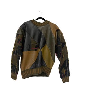 May include: A vintage sweater with a unique patchwork design. The sweater features a combination of leather and knit fabrics in shades of brown, black, and olive green. The sleeves have a patterned design with accents of pink and black.