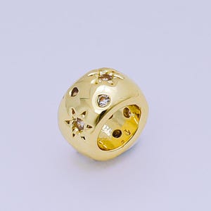 May include: A gold-colored charm bead with star-shaped cutouts and small, clear gemstones. The bead has a rounded, cube-like shape and is designed for jewelry making. The charm is shown against a light background.