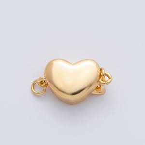 May include: A gold-colored heart-shaped clasp. The clasp has a smooth, polished surface and is connected to two small loops on either side. The heart is centered in the image against a white background.