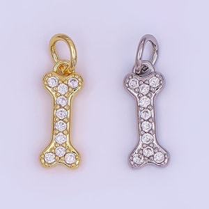 May include: Two dog bone-shaped charms. One is gold-toned, the other silver-toned. Both charms are encrusted with small, clear crystals. Each charm has a small loop at the top for attaching to a necklace or bracelet. The background is a light, solid color.