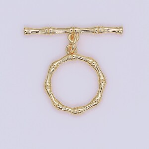 May include: A gold-coloured toggle clasp with a bamboo-style design. The clasp comprises a circular ring and a horizontal bar, both with textured details. The jewellery component is set against a light purple background.