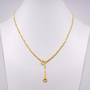Puede incluir: Un collar dorado exhibido en un maniquí blanco. El collar presenta un delicado diseño de cadena con un colgante de barra vertical y un pequeño cierre. El collar es de color oro brillante.