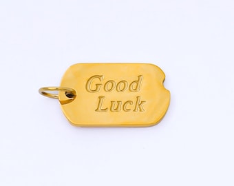 May include: A gold-colored rectangular pendant with rounded edges and a small loop. The pendant has the words "Good Luck" engraved on it. The background is plain white.