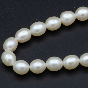 May include: A strand of creamy white, oval-shaped pearls against a dark background. The pearls are strung together, creating a necklace or bracelet. The pearls have a smooth, lustrous surface, reflecting light.