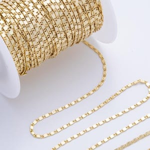 May include: A spool of gold-coloured chain with a flat, rectangular link design. The chain is partially unraveled, showing the pattern. The spool is white, and the background is a plain white surface.