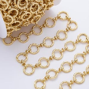 May include: A gold-toned metal chain composed of interconnected circular links with a textured, twisted surface pattern. The chain is wrapped around a white plastic spool, with a portion of the chain extending outward in a loose, folded arrangement.