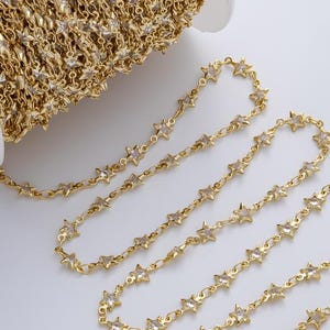 May include: A spool of gold-colored chain with star-shaped links. The chain is wound around a white plastic spool. Some of the chain is unwound and lies flat on a white surface. The stars have small, clear accents.