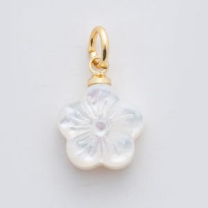 Puede incluir: Un colgante en forma de flor blanca con una argolla dorada. El colgante tiene reflejos iridiscentes, similares a la nácar. La flor tiene cinco pétalos y un centro detallado. La argolla es un simple lazo para sujetarlo a una cadena o pulsera.
