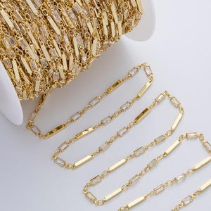 May include: A spool of gold-colored chain with rectangular links. The chain is partially unraveled, revealing its design. The chain is on a white spool, and the background is white.
