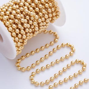 May include: A spool of gold-colored ball chain. The chain is made up of small, evenly spaced spheres. A portion of the chain is unraveled, lying in a curved line. The spool is white and has a central hole.